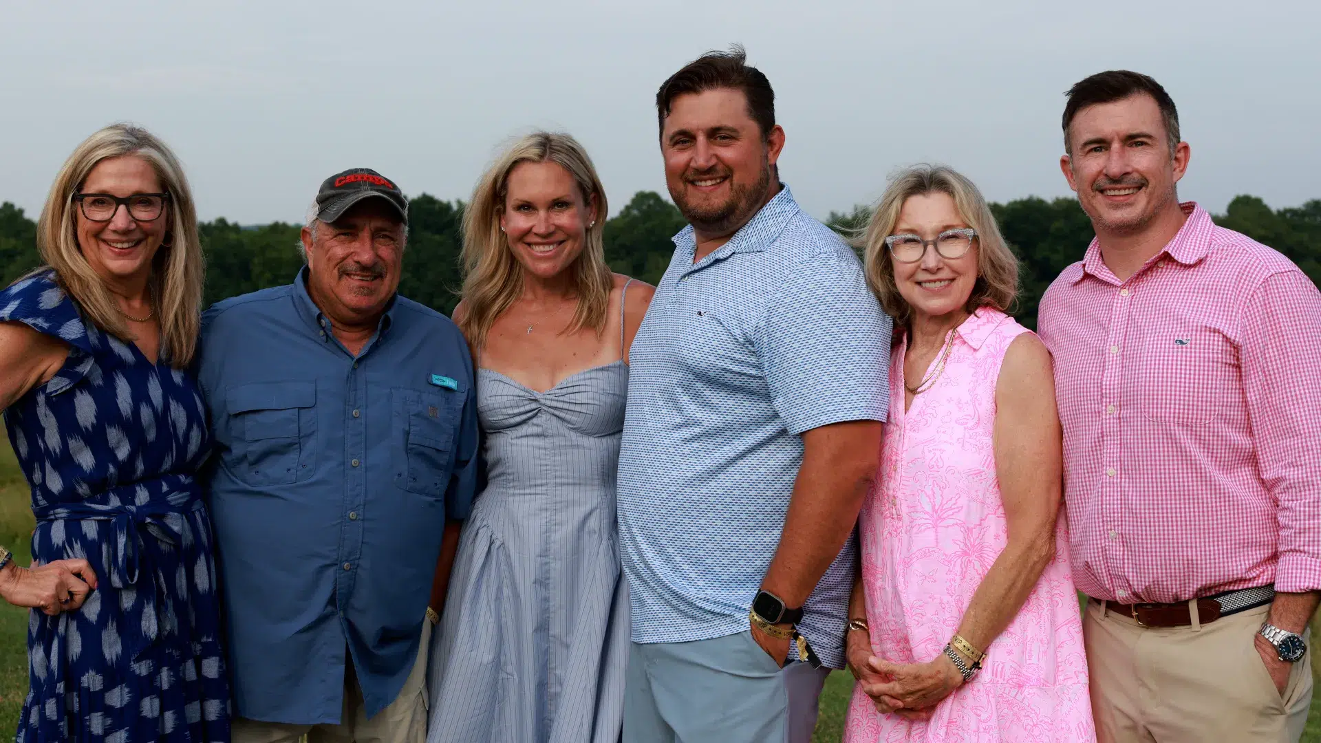 Six adults, dressed in casual wear, stand together in an outdoor setting with a light sky and distant greenery.